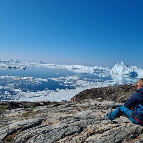 En kvinde i blåt vandretøj sidder på en klippe og kigger ud over isfjorden ved Ilulissat.