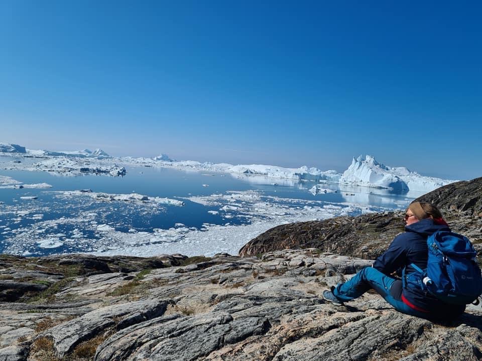 En kvinde i blåt vandretøj sidder på en klippe og kigger ud over isfjorden ved Ilulissat.