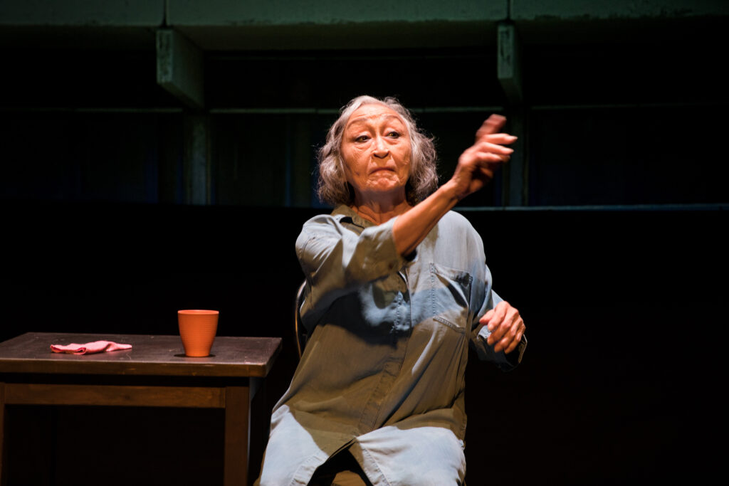 En kvinde, som sidder ved et bord alene på en teaterscene peger ud mod publikum.