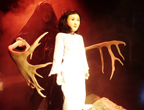 En dukke klædt i hvid omringet af gevir i et mørkt belyst scene