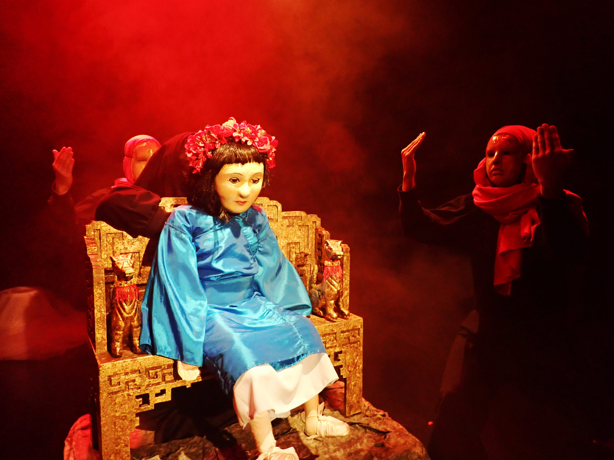 En dukke på en trone, med monke ved hendes side i et mørkt belyst scene